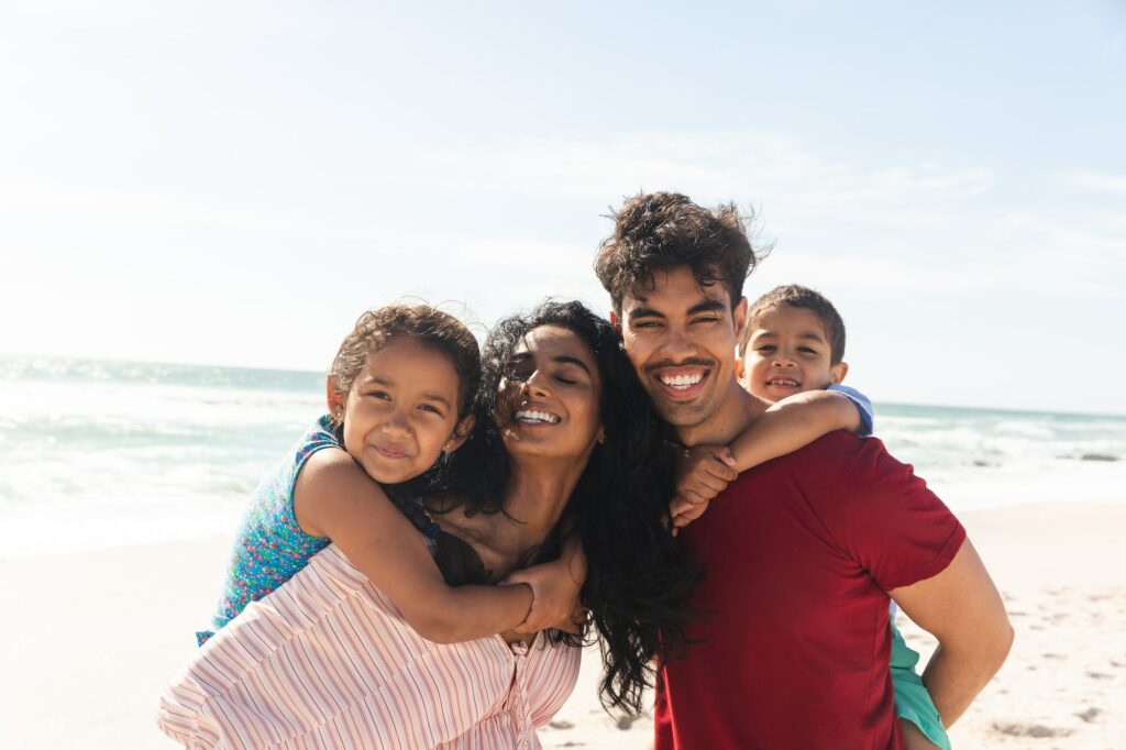 Happy biracial father and mother giving piggyback rides to kids at beach against sky with copy space